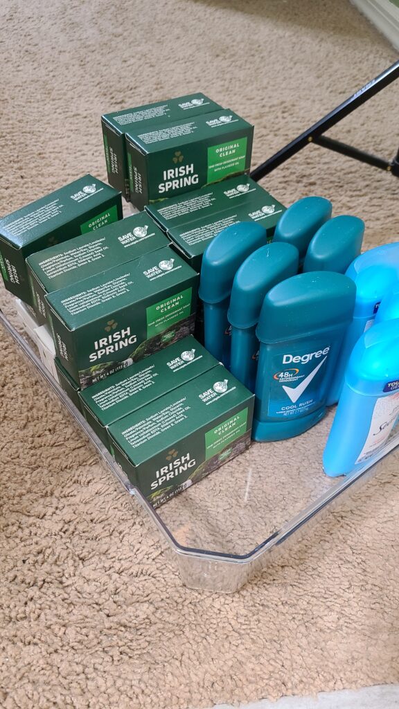 A clear storage bin sitting on the floor. It's filled with bar soap and deodorant.