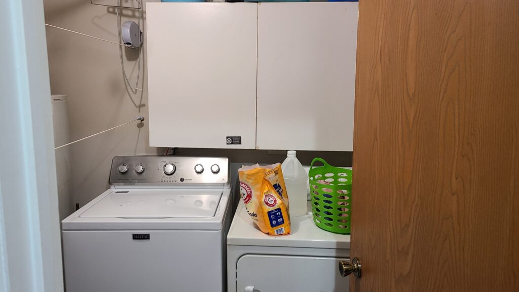 An open door leading to a small laundry room. A washer and dryer are in front, with closed cabinets hanging on the wall.