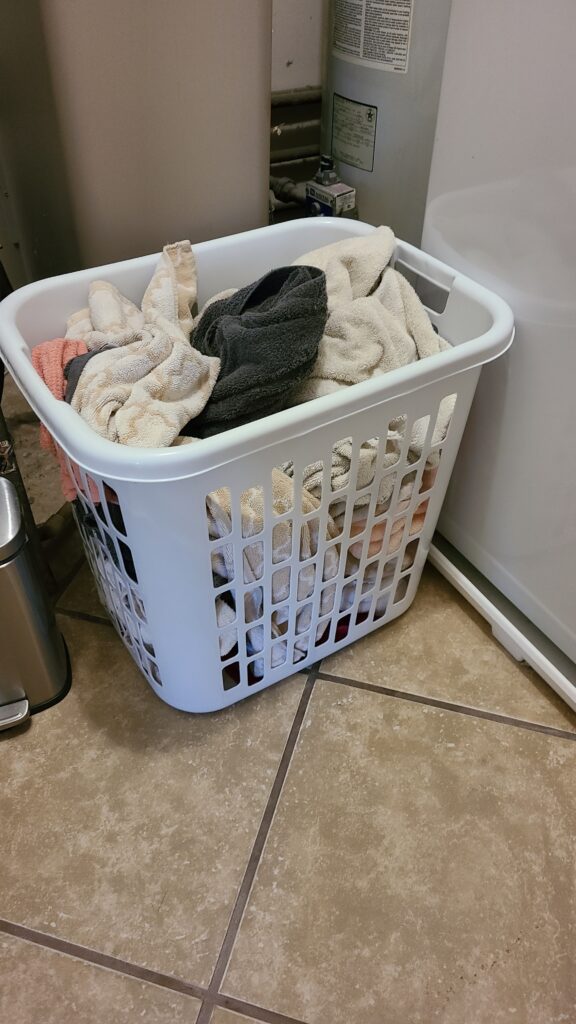 A tall white laundry basket in the laundry room. It is filled with dirty towels.