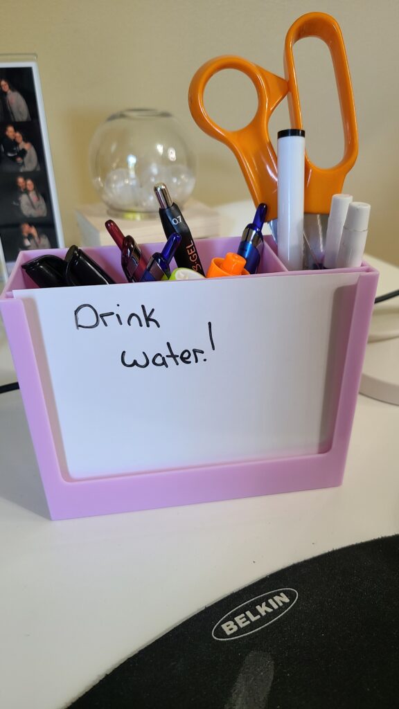 A pink and white pencil holder with various pens, markers, and scissors. There is a whiteboard attached to the pencil holder with the words "Drink water" drawn on it.