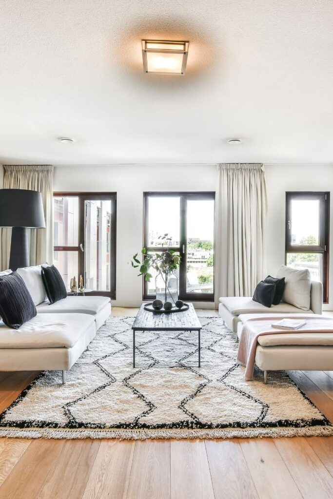 A bright and clean living room. Wooden floors and a white and black rug. Wide white couches with pillows on top. A coffee table sits between the couches, a glass flower vase on top.