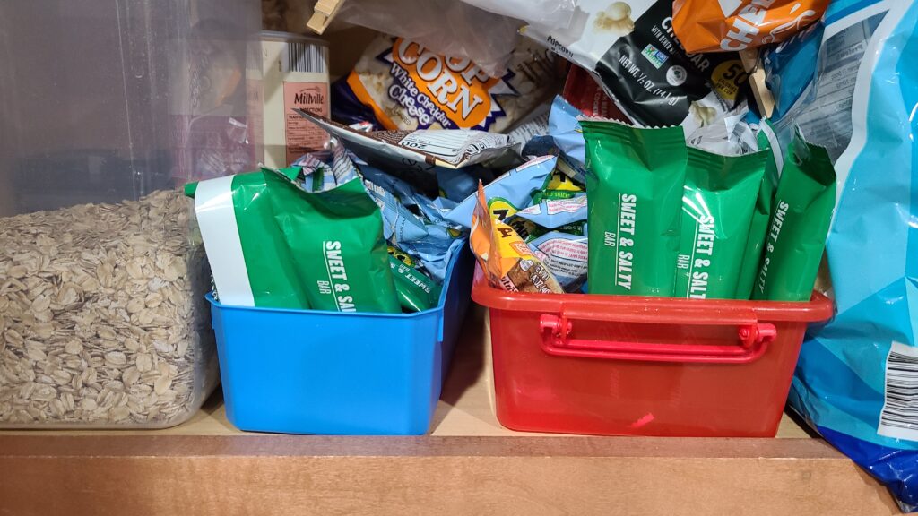 An open cabinet in the kitchen. Inside are two storage bins filled with snack bars and other snacks.