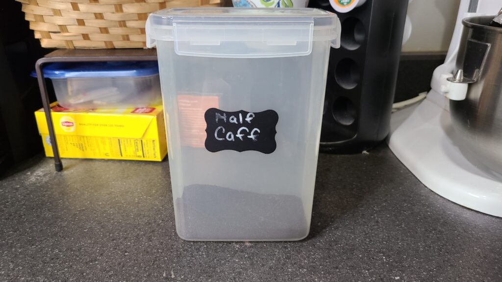 A food storage container filled with ground coffee. A label on the container says "Half caff"