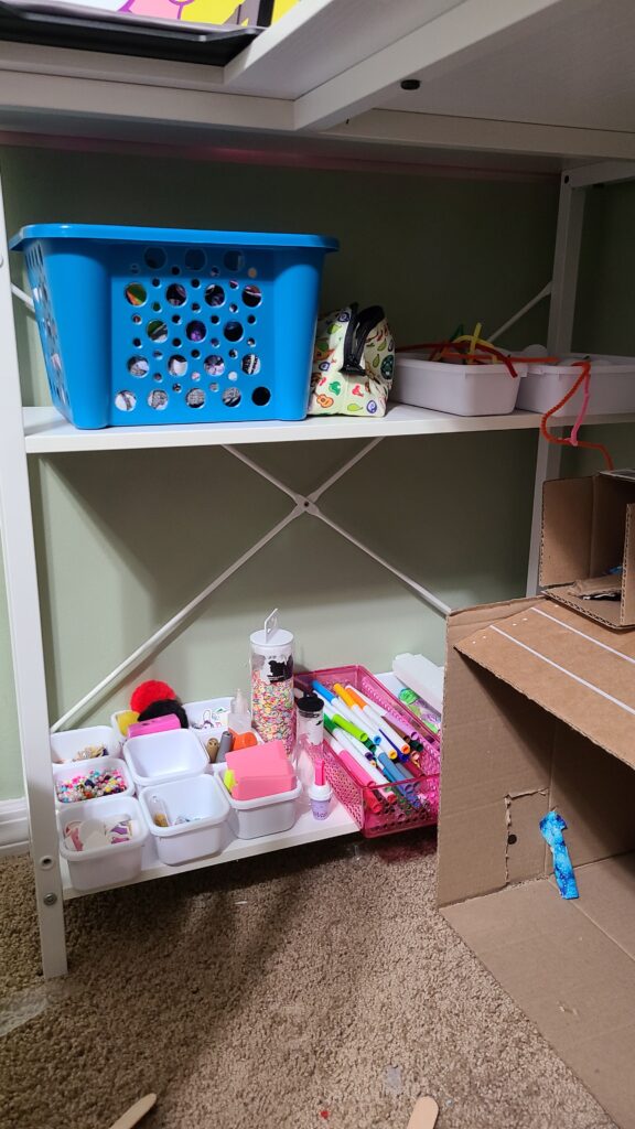 The underside of a desk, with two shelves. On them are drawer organizers filled with craft supplies.