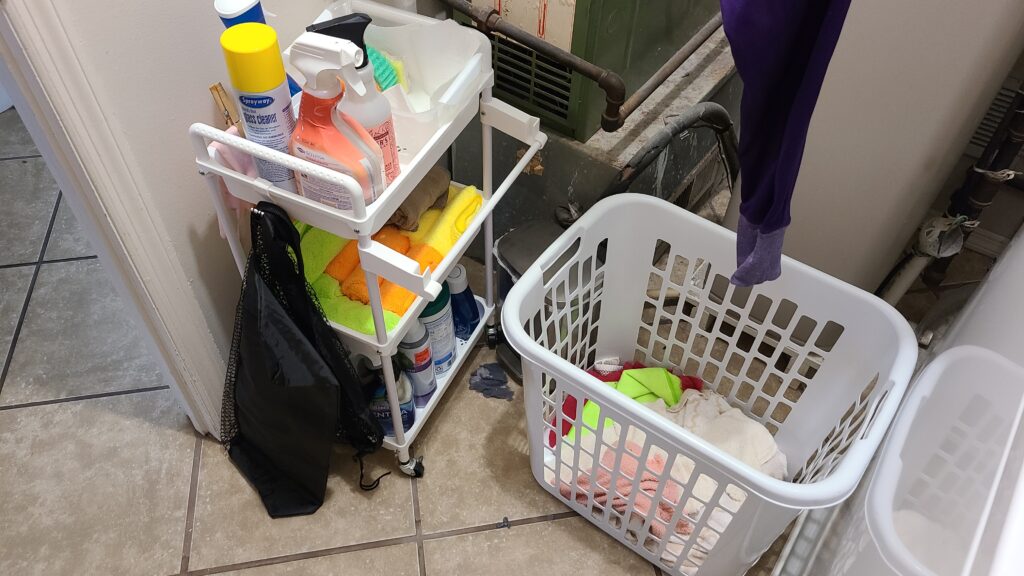 A small laundry room. In the photo are a rolling cart filled with cleaning supplies, and a laundry basket filled with towels.