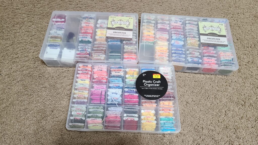 Three plastic craft organizers on a carpeted floor. Inside are bobbins of colored floss.