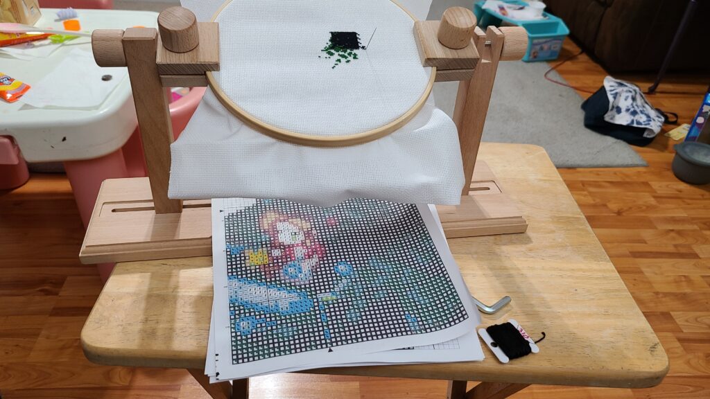 A small wooden folding lap table. On top is a wooden lap holder for a cross stitch.