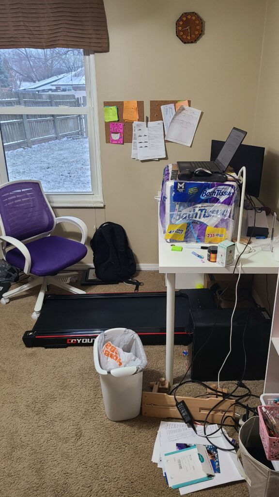 A desk in a home office. A walking pad is underneath, and a laptop is propped up on the desk with a package of toilet paper.